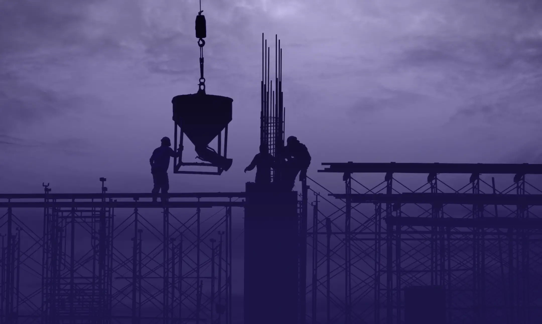 Silhouetted construction workers operate a crane to lift a concrete bucket atop a high-rise structure, set against a dusky, purple-hued sky. Scaffolding and rebar frames are visible, highlighting the about us section of this construction scene against the dramatic evening backdrop.