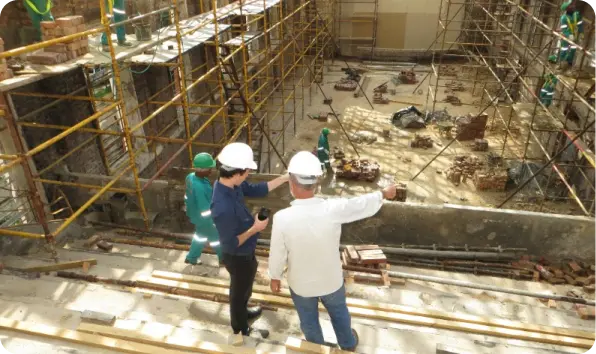 Two construction workers in white hard hats stand on a scaffolding platform, overlooking a building site with scaffolding on both sides and workers in green uniforms working below. One points towards a specific area while the other holds a roll of plans. Learn more about us as we shape the future.