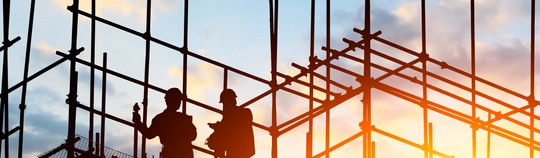 Silhouettes of two construction workers standing on scaffolding against a sunset sky. The workers appear to be discussing something as they hold tools. The intricate framework of the scaffolding creates geometric patterns against the warm, glowing sky, embodying the essence of our "About Us" story in every angle.