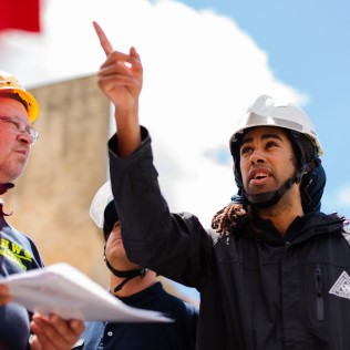 A man in a black jacket and white helmet points upward while speaking to two other men, also wearing helmets. One of the other men, in a yellow helmet and glasses, holds papers. The sky in the background is bright with scattered clouds.