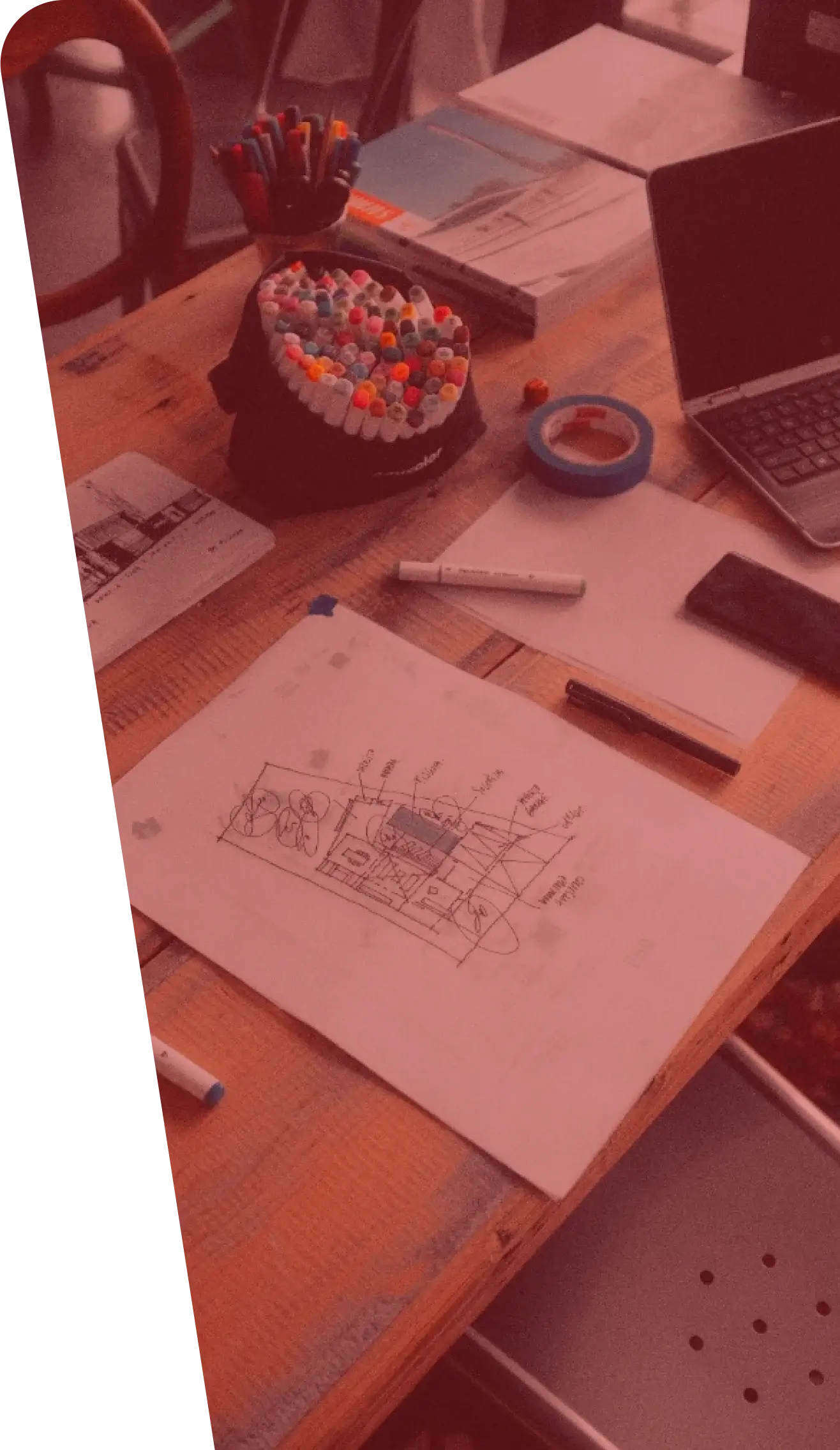 A wooden desk cluttered with various items including a laptop, numerous colored markers, a technical drawing, open books, a black wallet, and a roll of blue tape resembles the workspace of someone preparing essential services. The technical drawing features detailed diagrams with annotations.