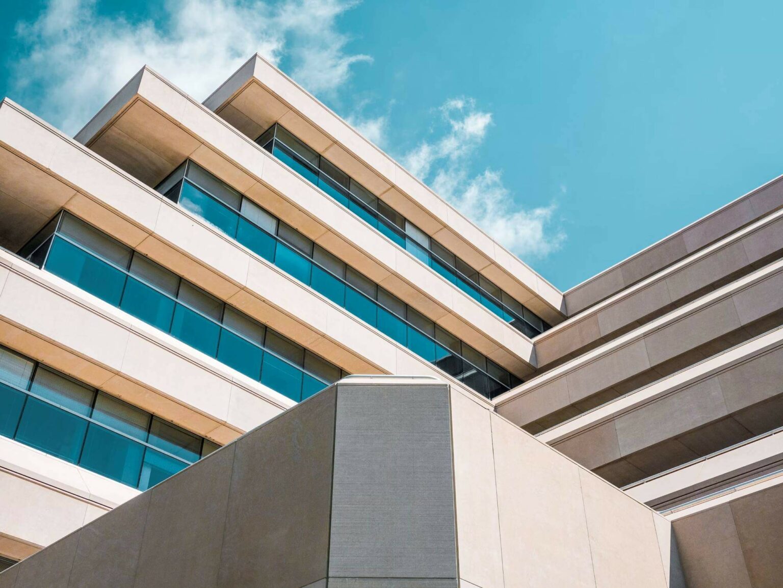 A modern, multi-story building with clean, geometric lines and large glass windows under a blue sky with a few scattered clouds. The facade features layered, staggered levels, creating an angular architectural design.