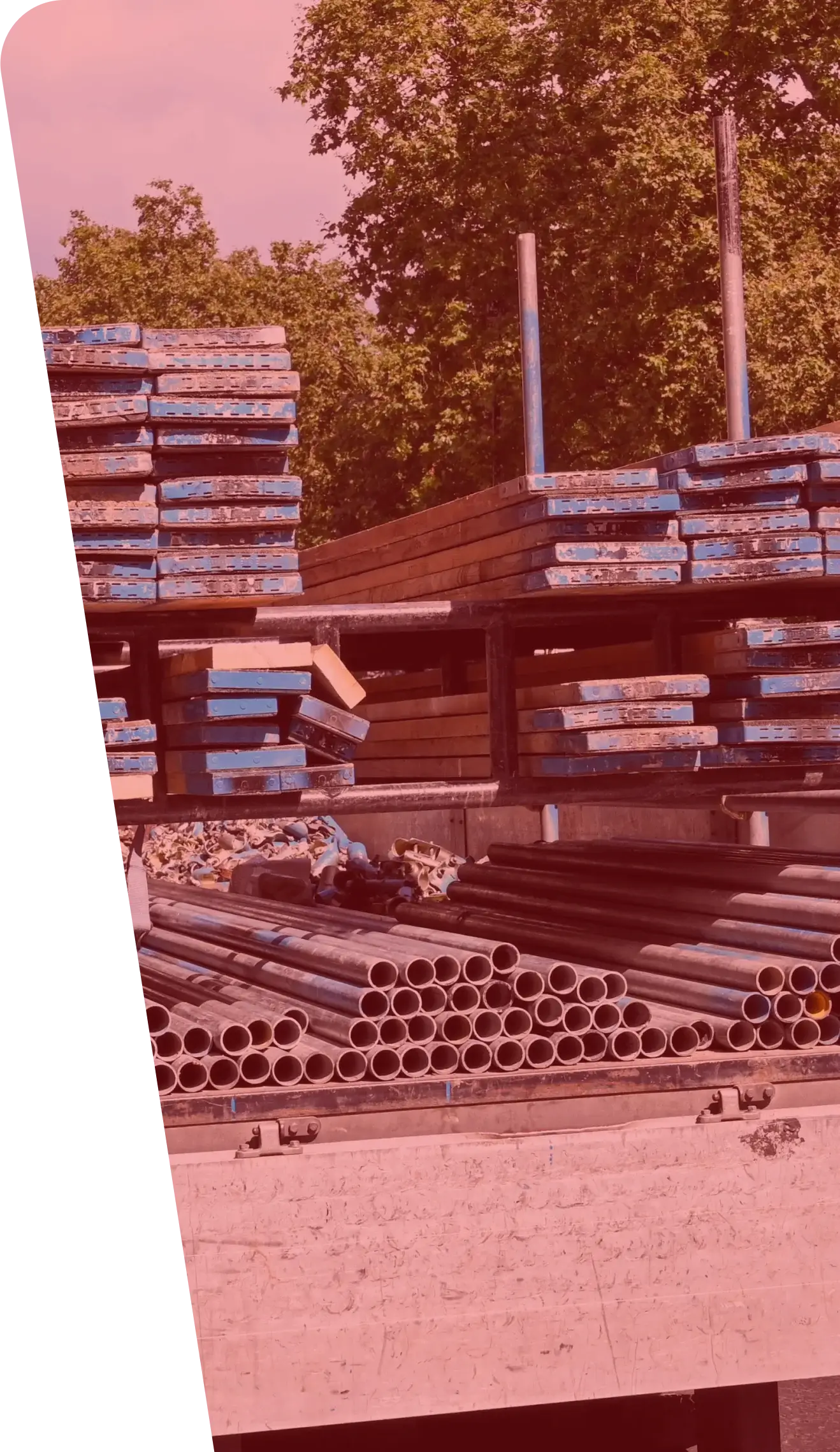 Stacks of metal pipes and rectangular wooden planks painted with blue and red colors are organized at a construction site. The materials, essential for construction services, are arranged in an orderly manner, and trees are visible in the background.
